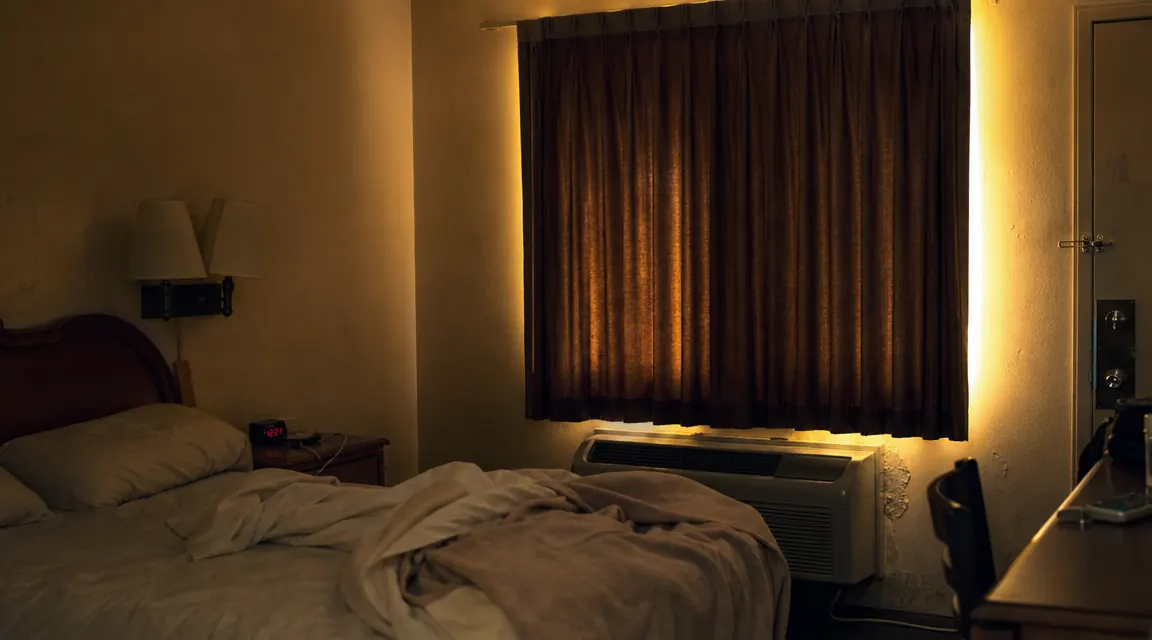 Worn motel room with thin curtains and bright parking lot light bleeding through the gap